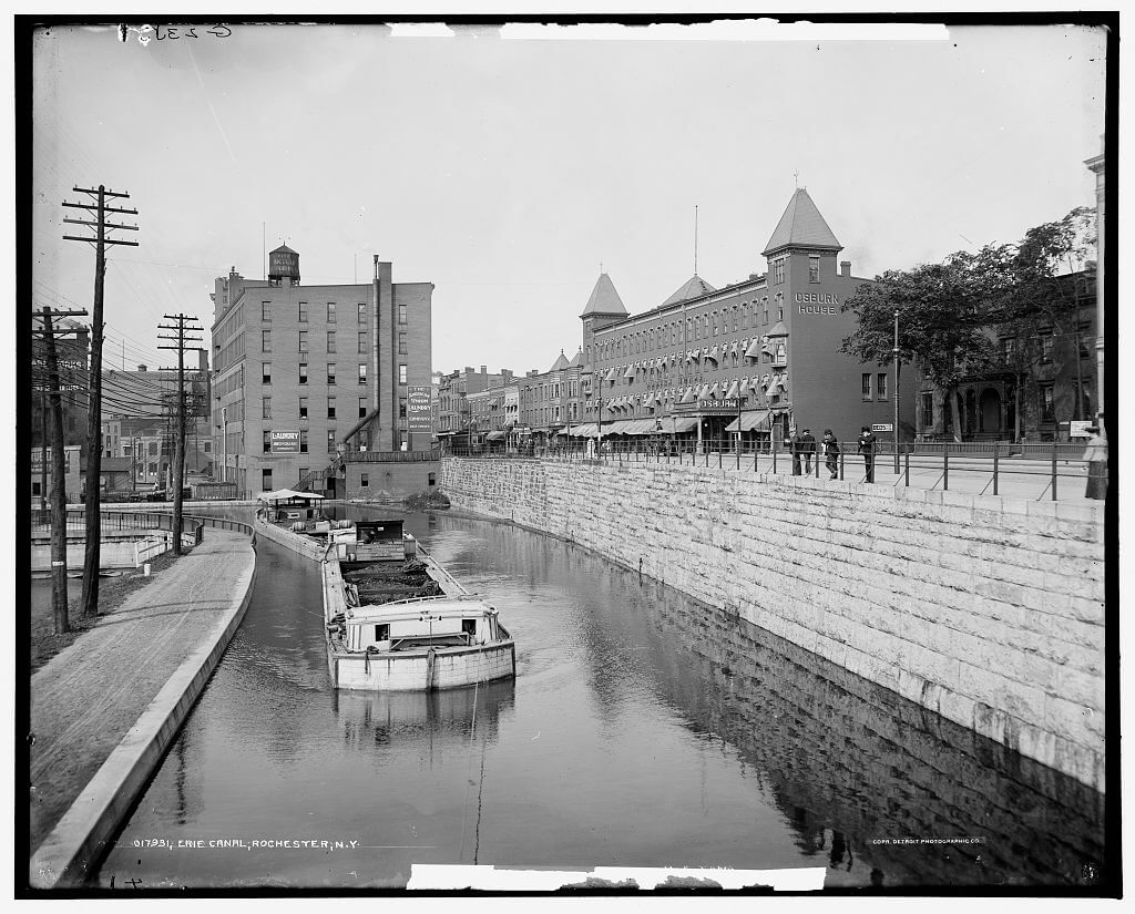 Canal boats moored along the Erie Canal in Rochester, New York, with buildings and bridges visible in the background, circa 1900–1906