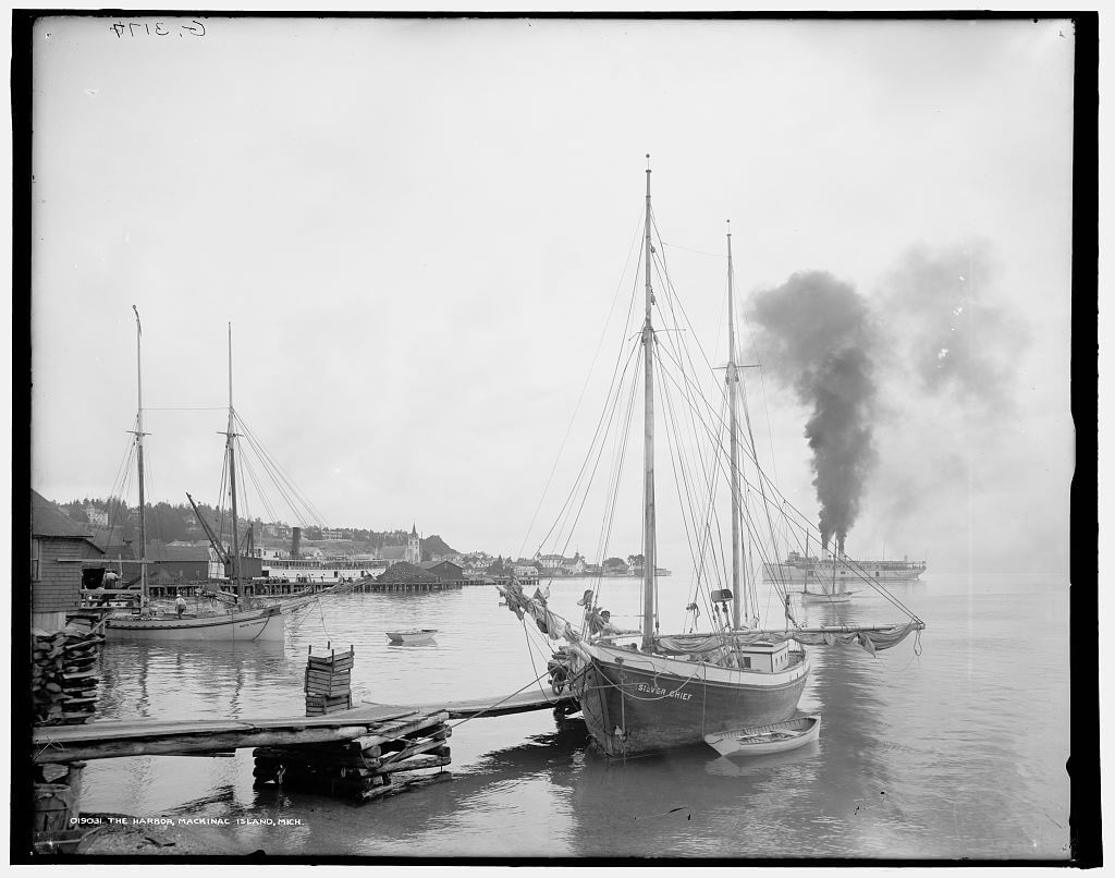 Two Great Lakes schooners moored in a harbor, their tall masts and rigging silhouetted, with a steamship visible in the background