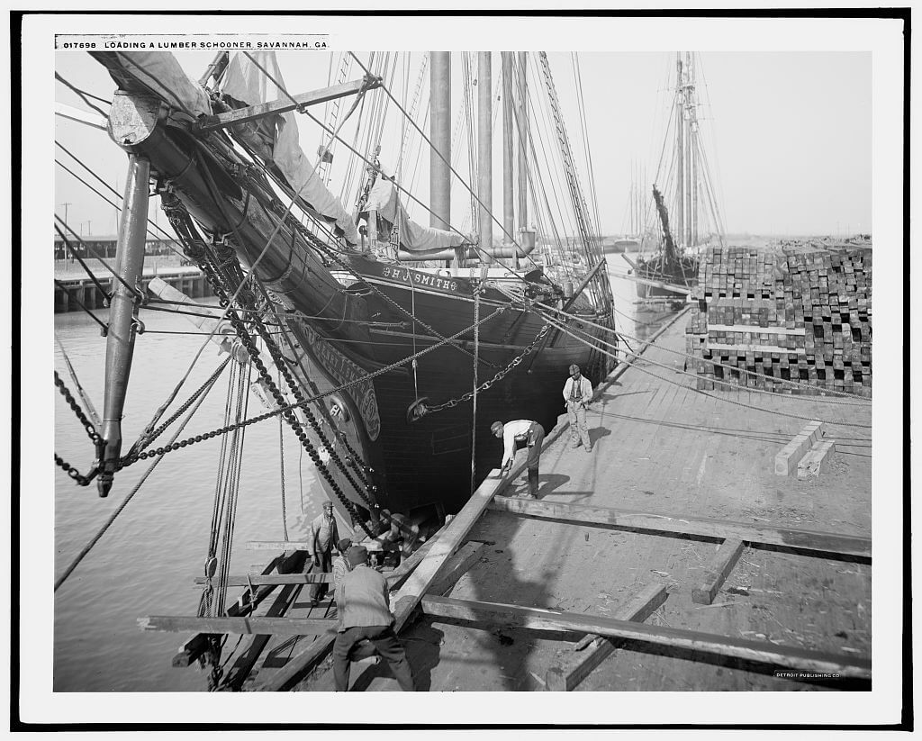 Workers loading stacked lumber onto a schooner at dock, showing the scale of the lumber trade with piles of boards stacked on the wharf, circa 1900–1906