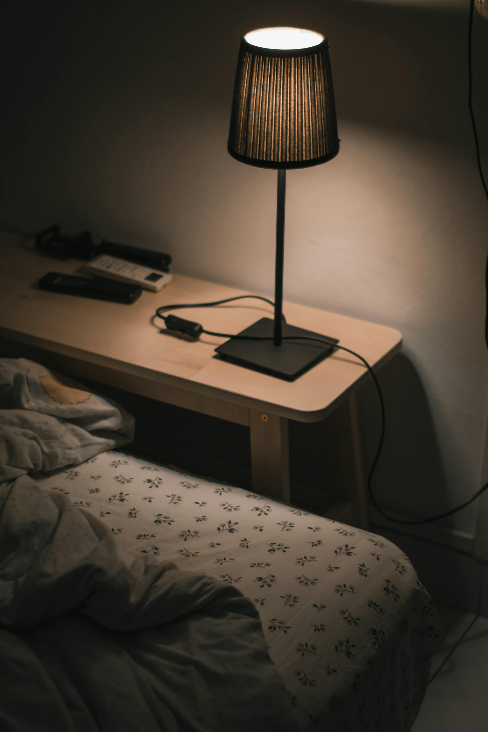 A small black bedside lamp casting a warm glow on a wooden nightstand next to a patterned bedspread in a dimly lit bedroom