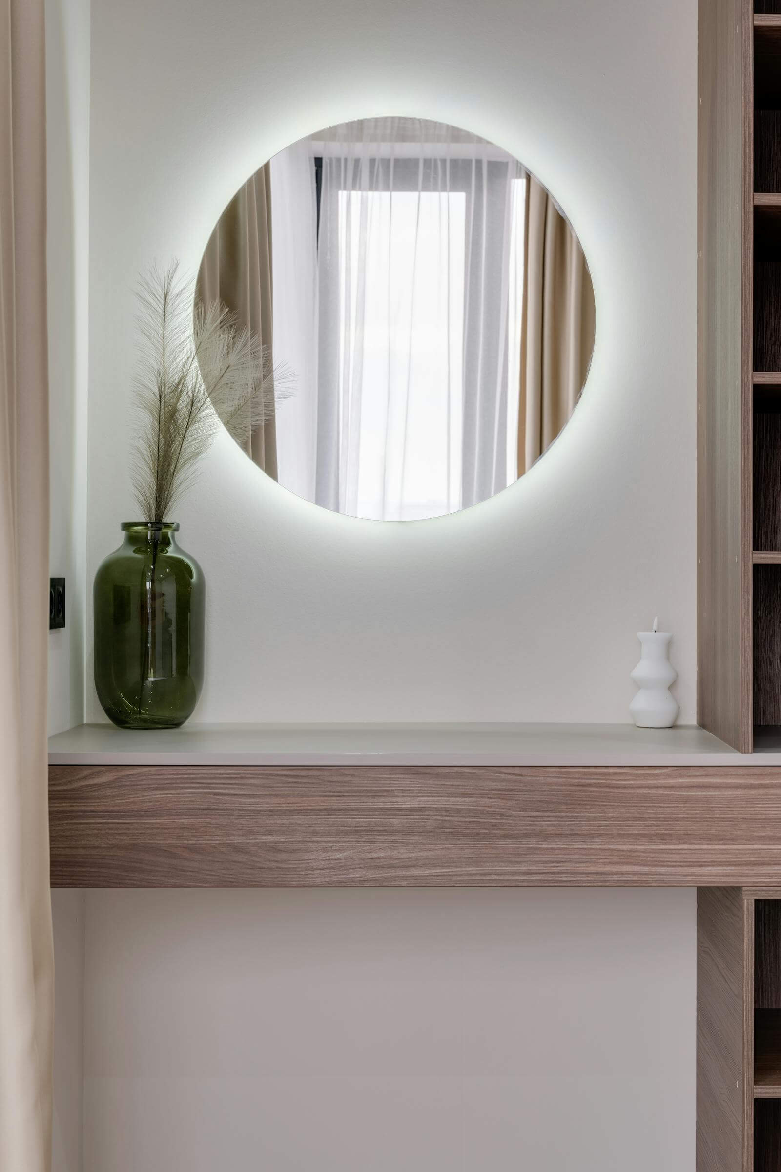 A round backlit mirror hanging above a slim wooden console in a small apartment, with a green vase of dried pampas grass and a white candle, reflecting light from a curtained window