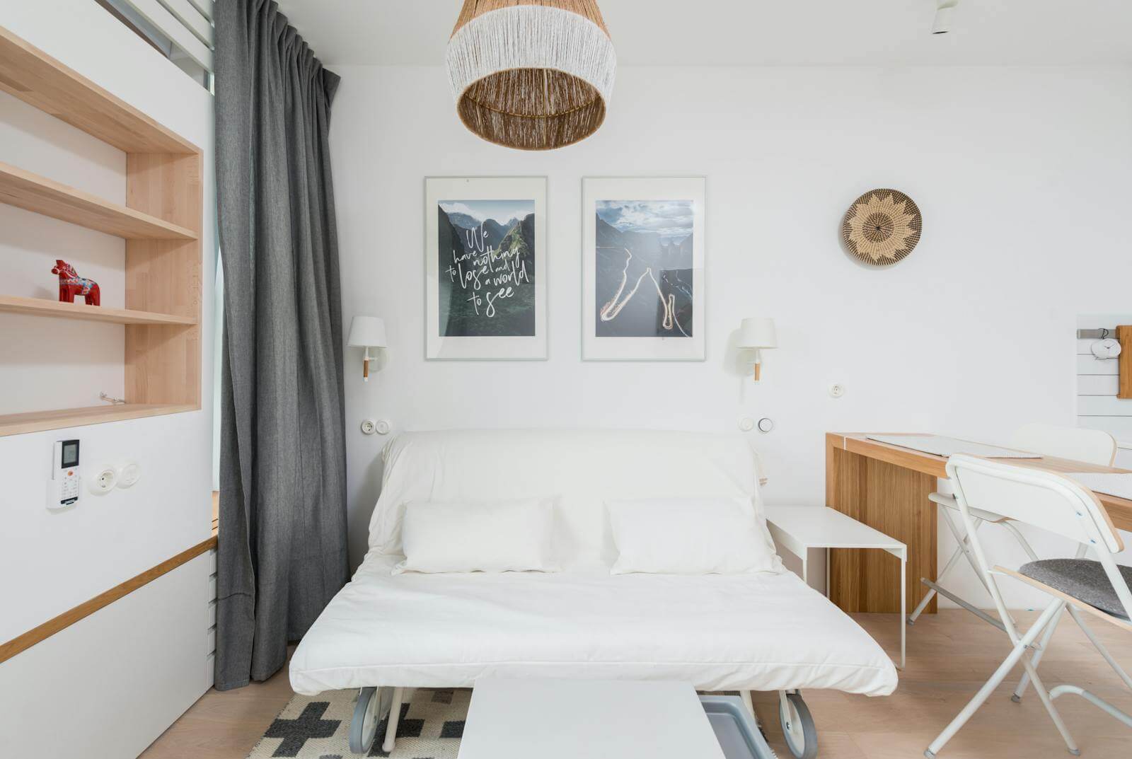 A white sofa bed pulled out flat into a mattress in a small studio apartment, with built-in wooden shelving on the left, a folding chair and slim desk on the right, and framed art on the wall above