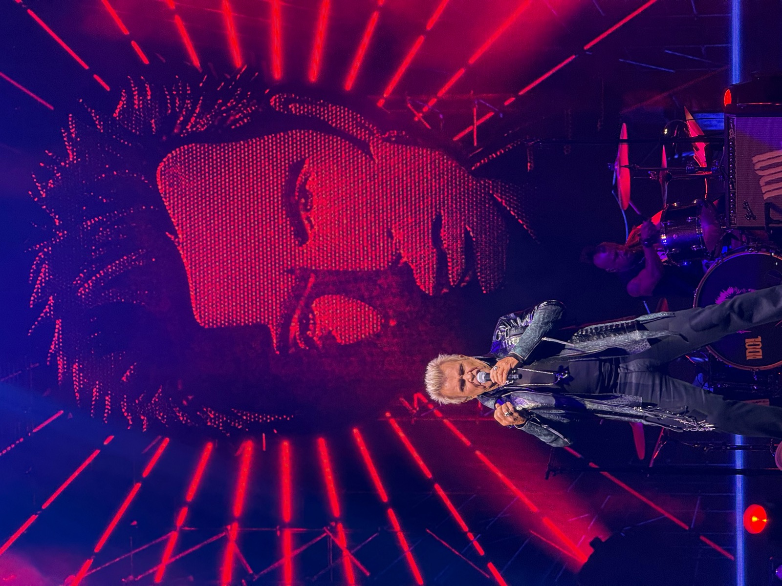 Billy Idol performing on stage in front of a giant LED screen showing his portrait