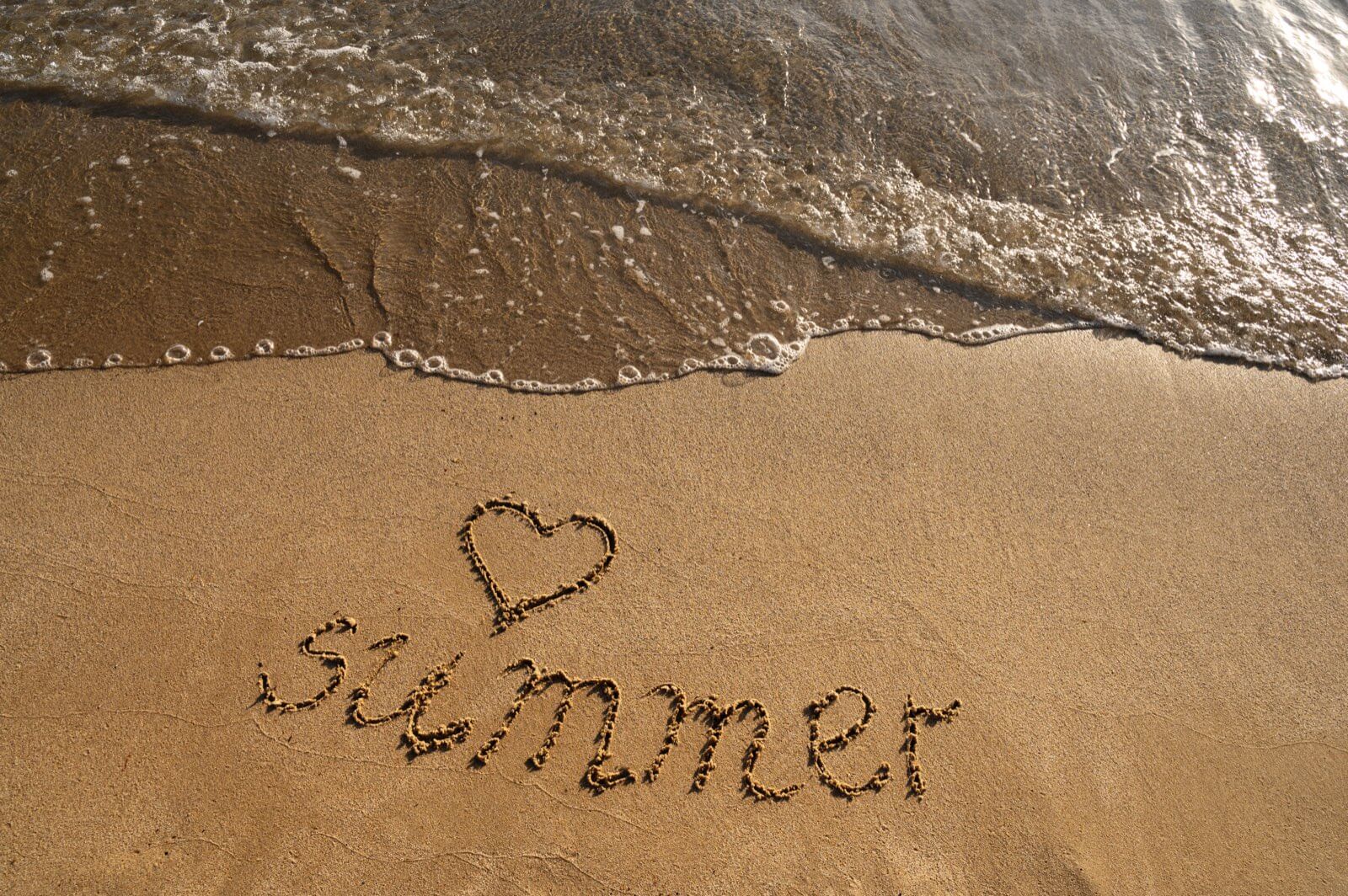 The word summer with a heart drawn in the wet sand as a wave washes up the beach