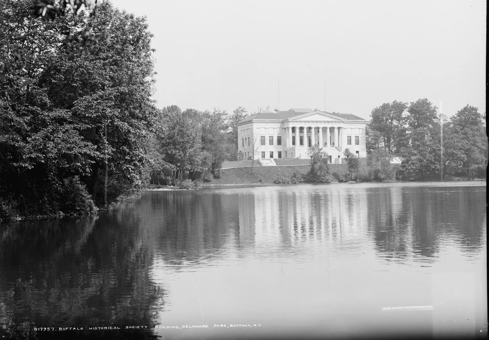 Black and white photograph from about 1904 of the Buffalo Historical Society building, a white marble Greek revival structure with a columned portico, seen across still water from the far bank of the lake