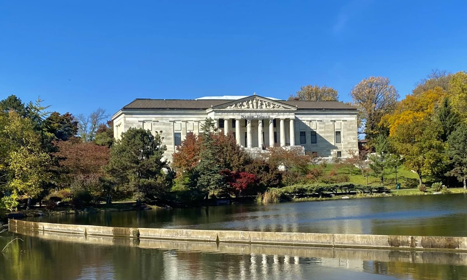 The Buffalo History Museum seen across Mirror Lake in autumn, its white marble columns rising above the Japanese Garden and trees in red and gold