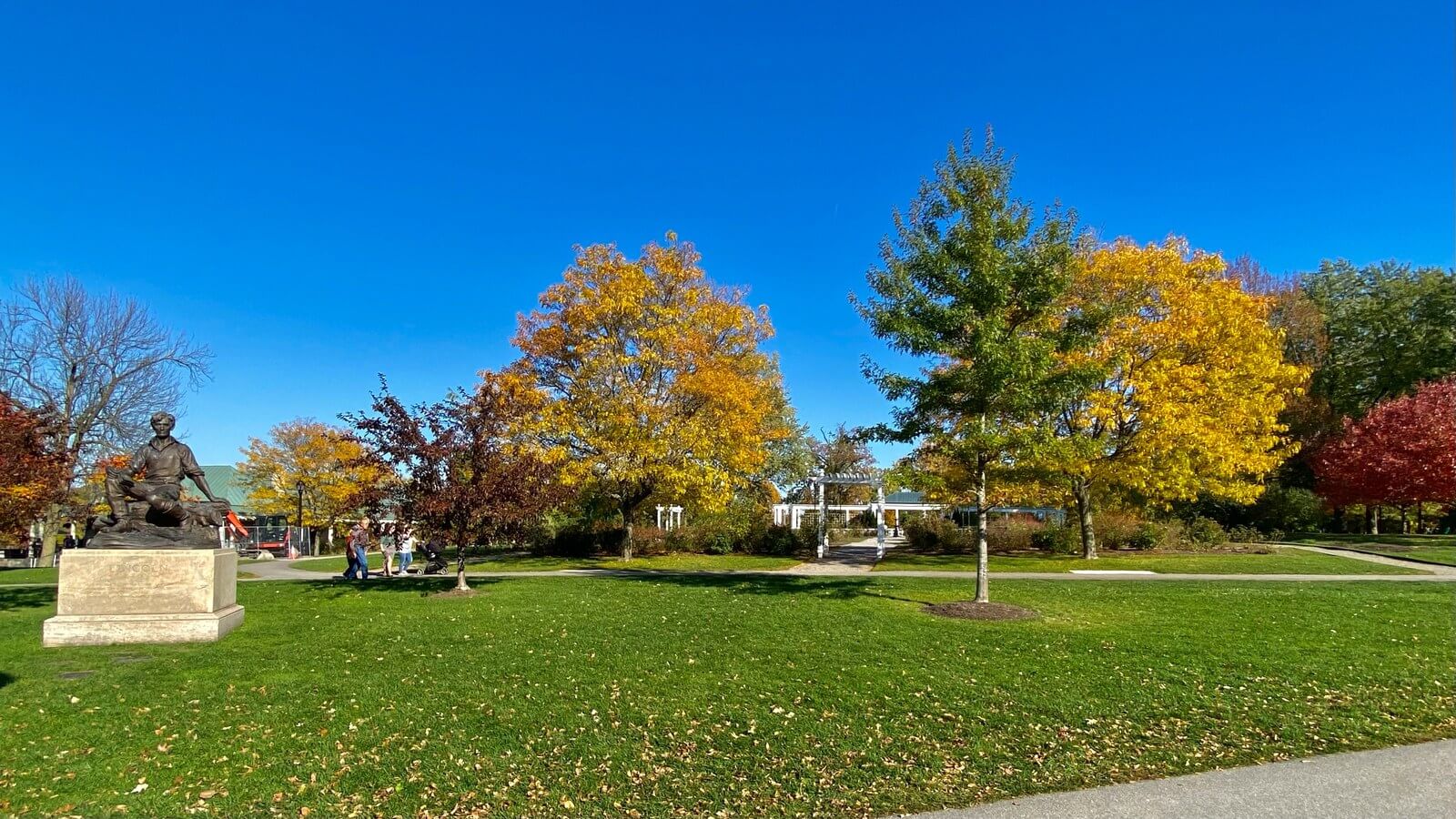 Trees in gold and orange autumn color above a green lawn in Delaware Park, with the Young Lincoln statue on its stone plinth at the left and a deep blue sky overhead