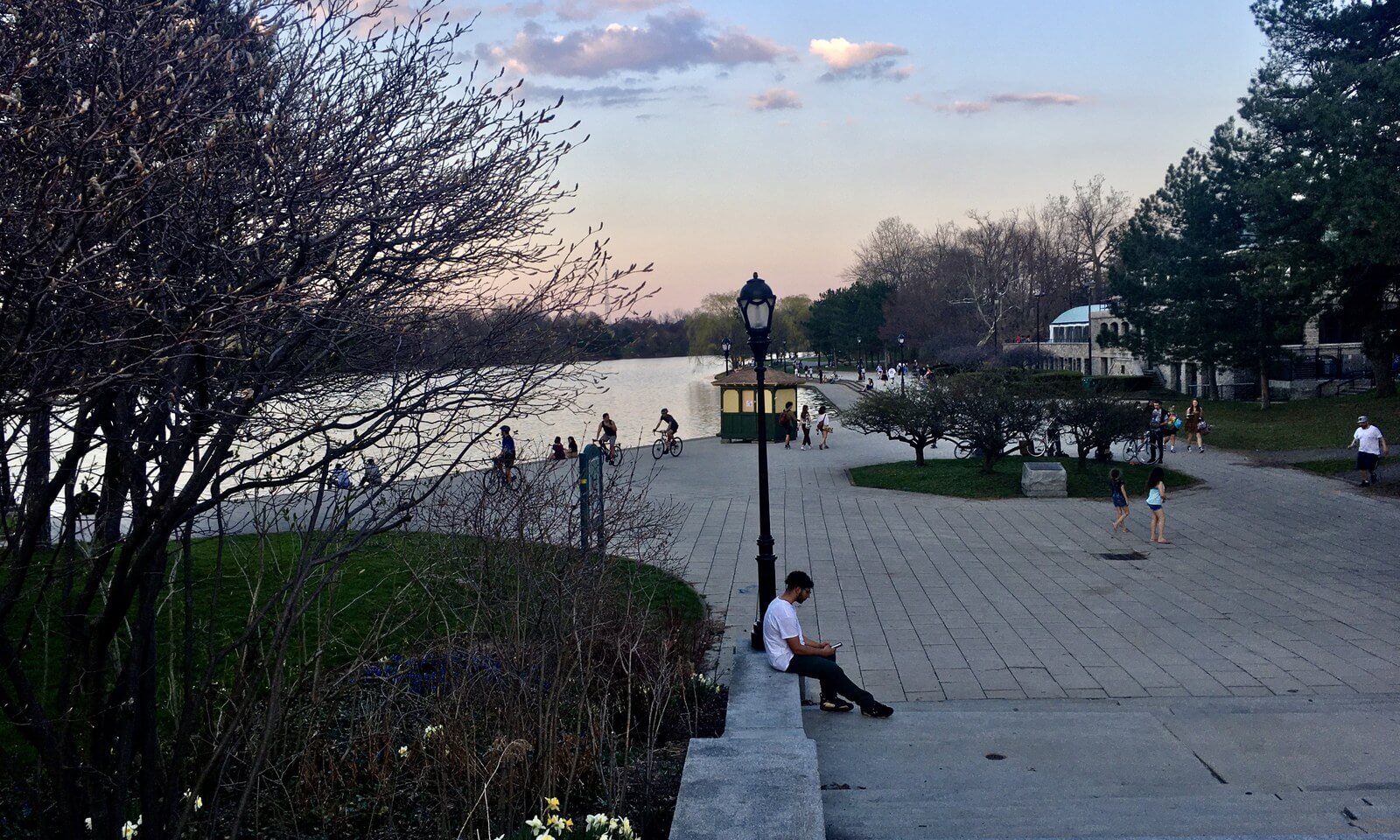 People walking and sitting along the paved terrace beside Hoyt Lake in Delaware Park at golden hour on an April evening, with lamp posts along the promenade and the trees still bare