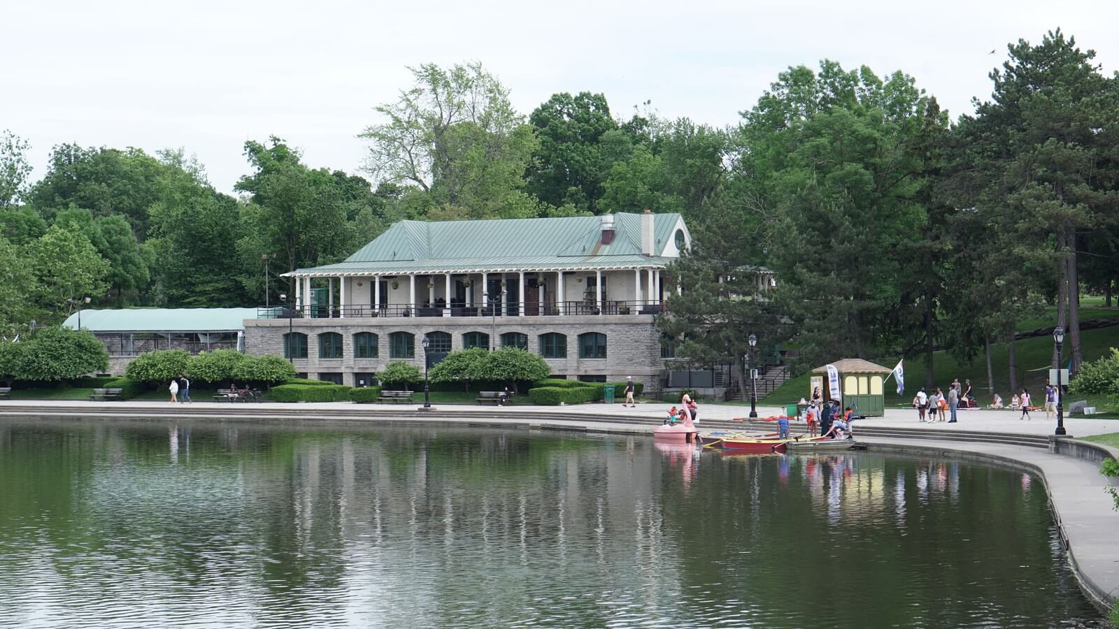 The white Marcy Casino building reflected in Hoyt Lake on a summer day, with people gathered along the shoreline path and small boats on the water