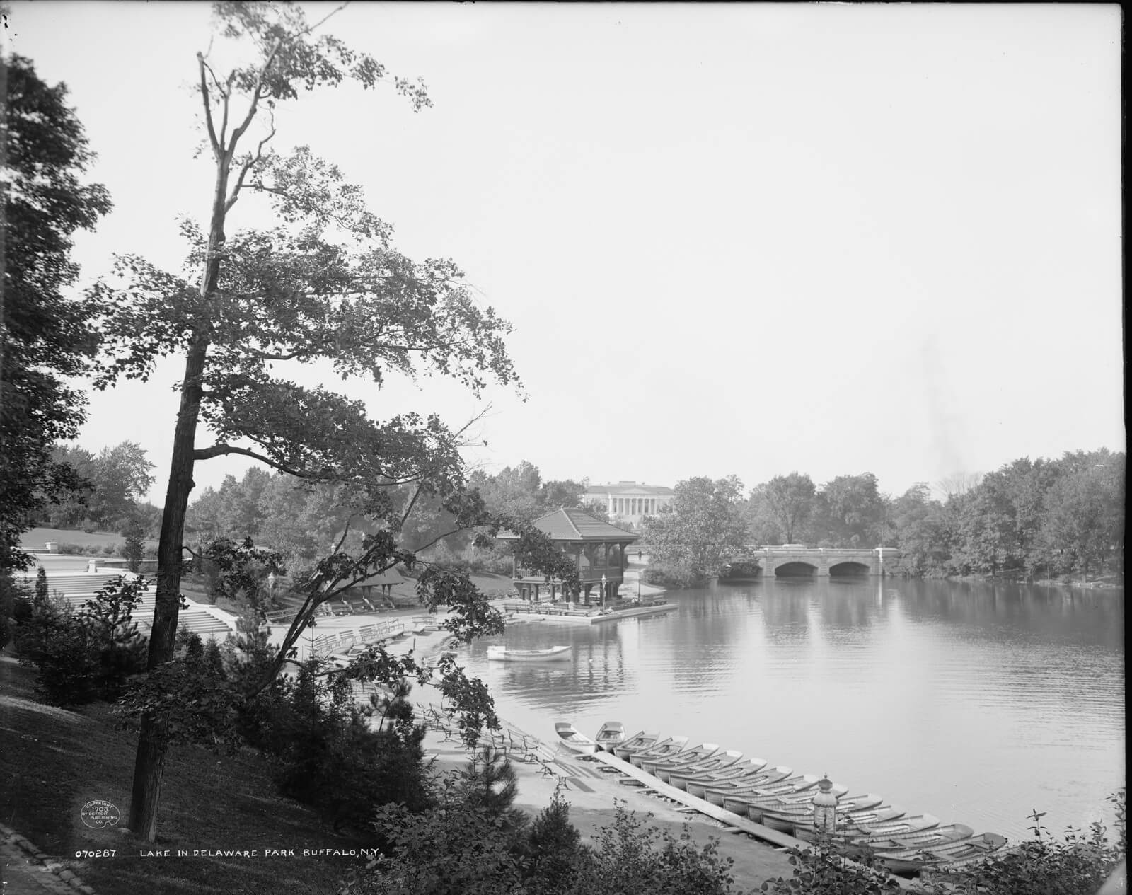 Black and white photograph of Hoyt Lake in Delaware Park around 1908, with a long line of rowboats moored at a dock in the foreground, a wooden boathouse pavilion on the shore, a stone arch bridge across the water, and the Historical Society building on the hill beyond