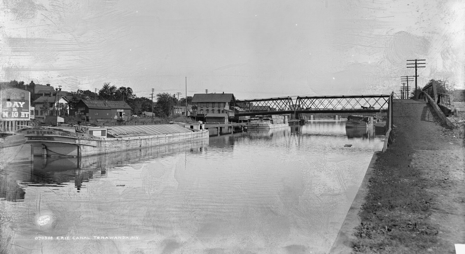 Black and white photograph of the Erie Canal at Tonawanda around 1900, with loaded canal boats moored along the left bank, an iron swing bridge crossing the water, and the stone canal wall and towpath running along the right