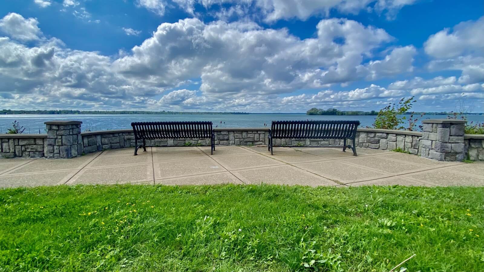 Two dark metal benches on a paved stone overlook facing the open water of the Niagara River, with green lawn in the foreground and tall white clouds above