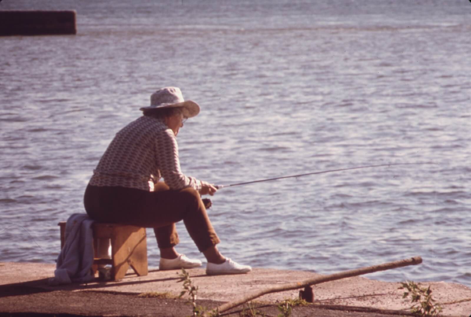 A lone angler in a wide-brimmed hat sitting on a low wooden stool at the concrete edge of the Niagara River, rod extended out over sunlit water