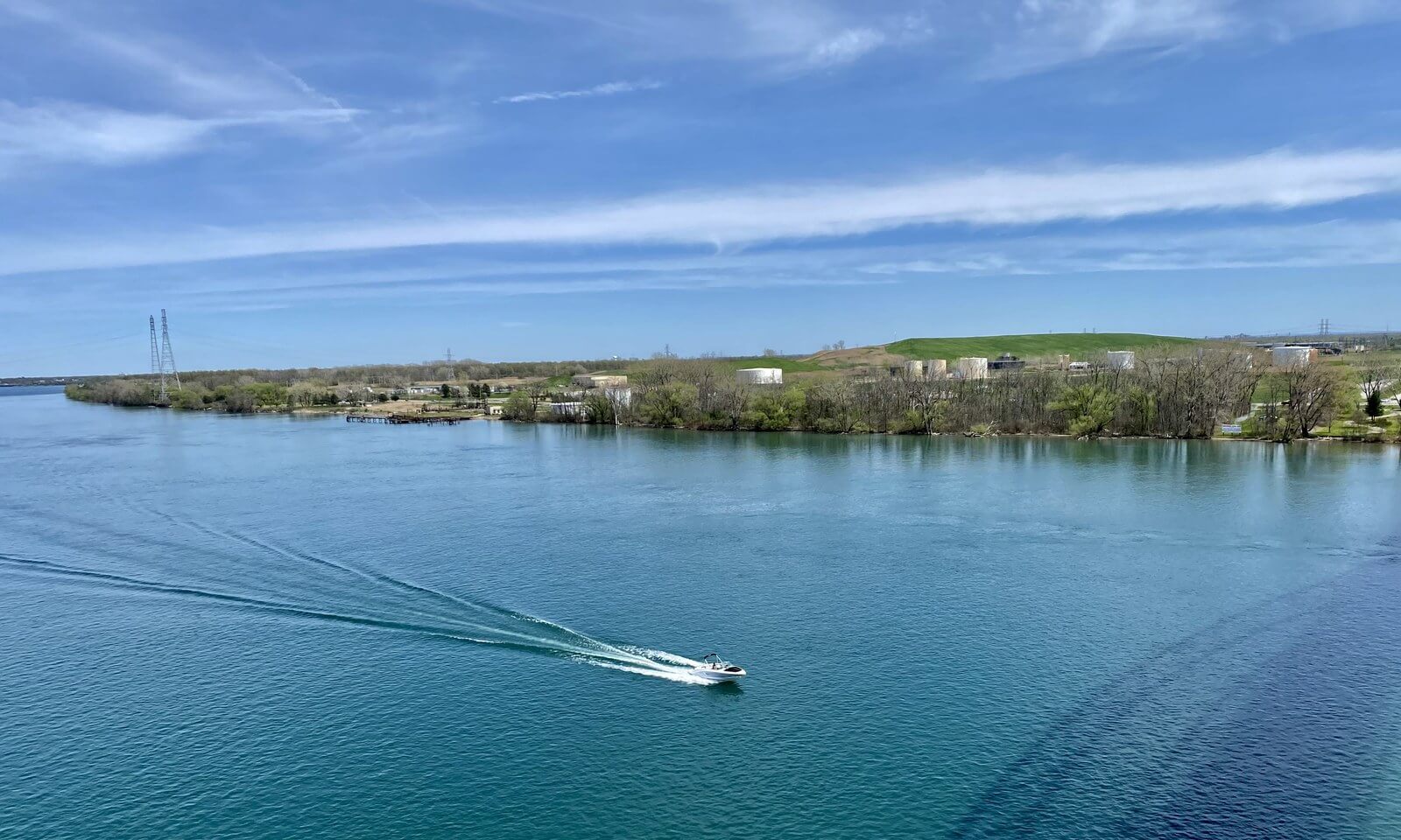 A small speedboat crossing the wide blue-green Niagara River near Tonawanda, leaving a long white wake behind it, with a wooded shoreline in the distance
