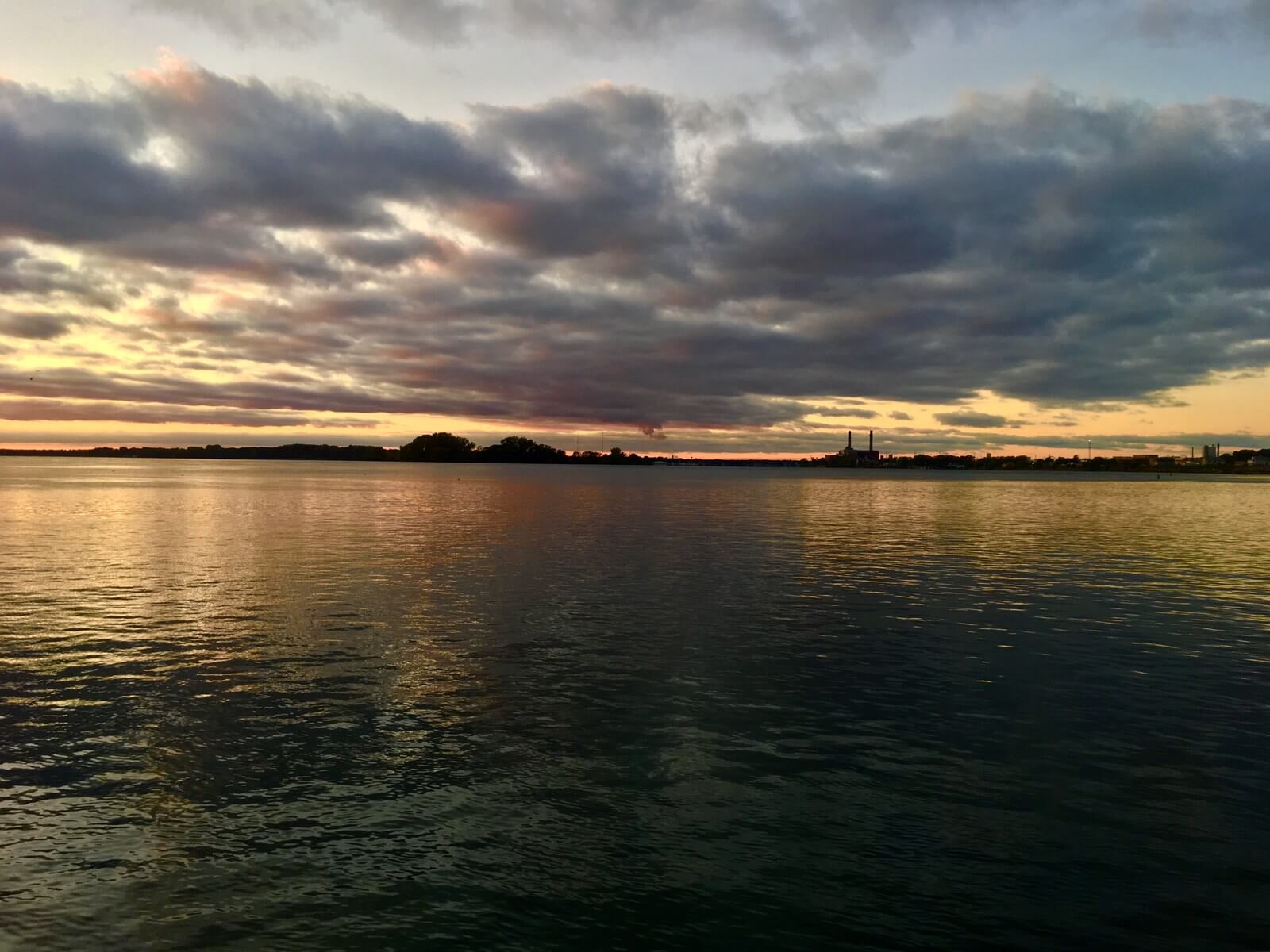 Sunset over the Niagara River with orange and gold light breaking beneath a bank of dark cloud and reflecting in a long streak across the dark water