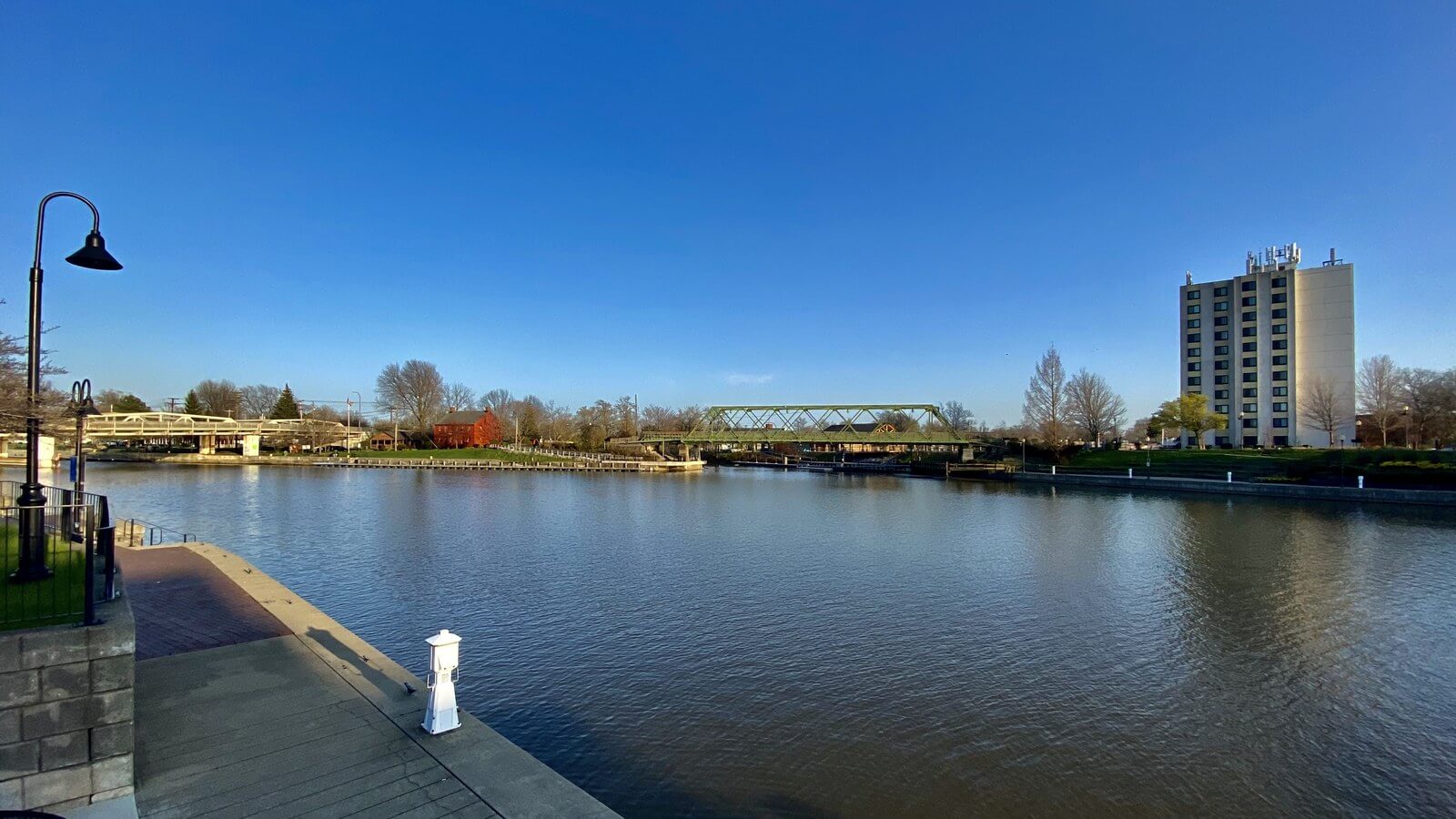 The paved waterfront promenade at Gateway Harbor on the Erie Canal between Tonawanda and North Tonawanda, with a lamp post, mooring bollard and open water under a deep blue sky