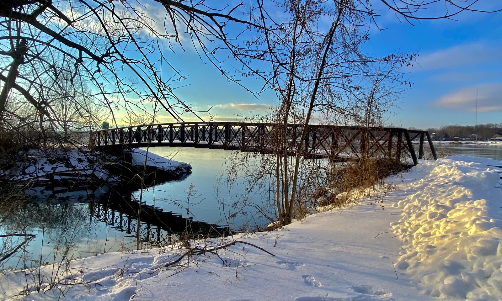 A steel footbridge crossing the snow-covered mouth of Two Mile Creek in Tonawanda at low winter sun, with bare branches overhead and ice along the bank