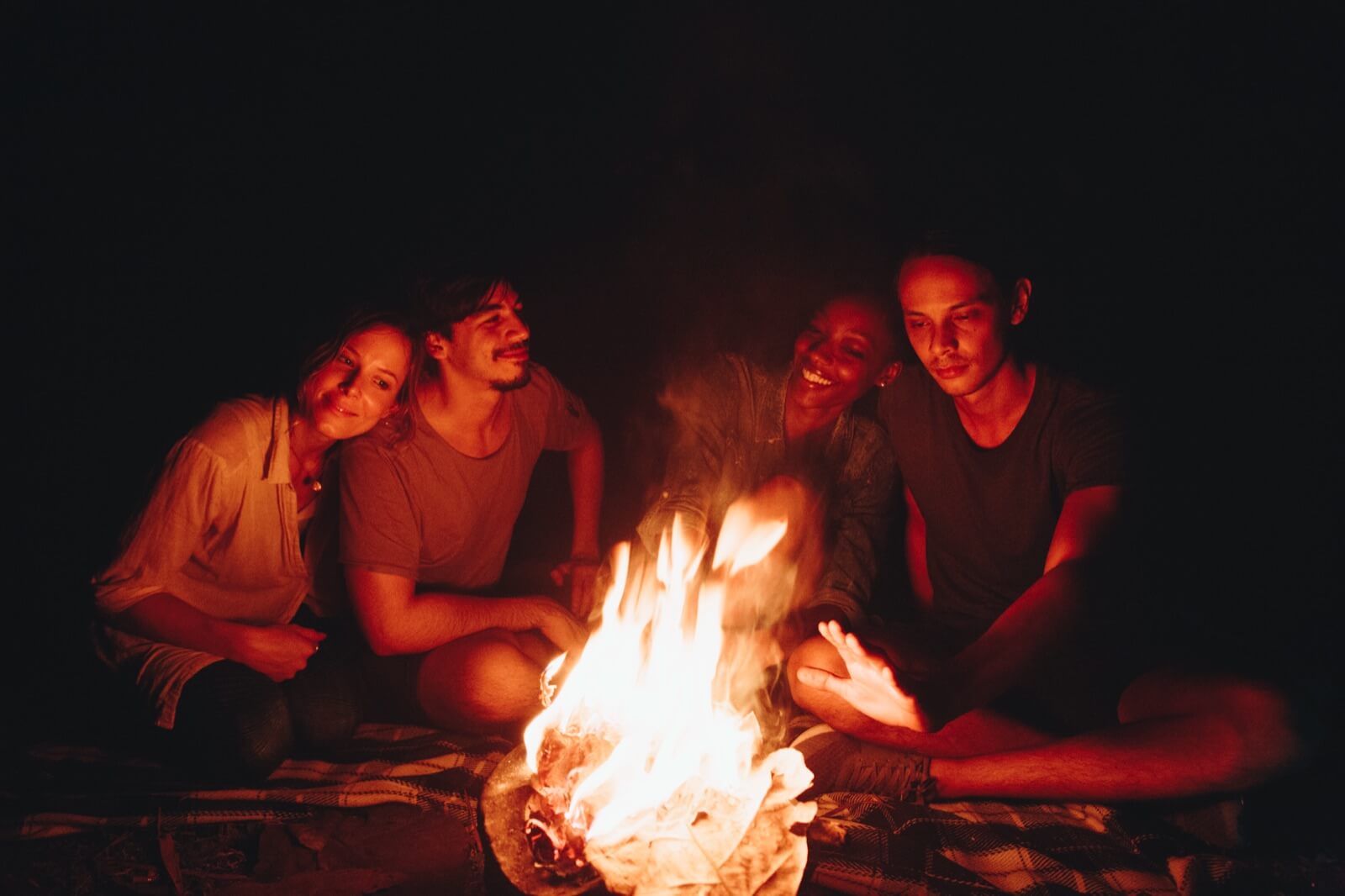 Four friends sitting close together around a crackling campfire at night, faces lit by the flames